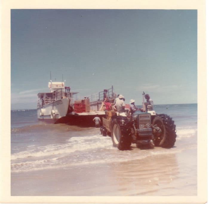 A tractor helps unload a barge on the beach in a 1970s photo.